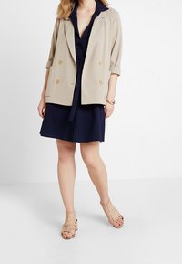 Béžový dvouřadý blejzr přes námořnické modré košilové šaty, doplněný béžovými sandály na tenkých řemíncích. Blejzr má ohrnuté rukávy a hladký povrch.