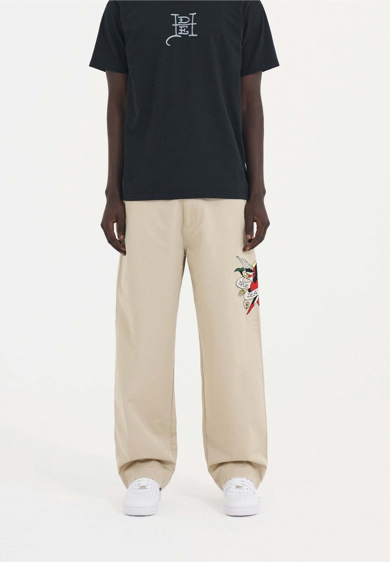 Personne portant un pantalon beige à jambes larges avec un motif coloré de style tatouage sur la jambe droite, associé à un t-shirt noir et des baskets blanches.