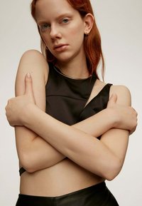 Top corto negro sin mangas con un diseño estructurado, que presenta un escote asimétrico y una textura suave. Los brazos del modelo están cruzados sobre el abdomen descubierto.