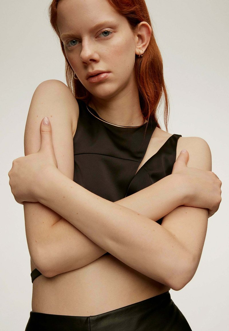 Top corto negro sin mangas con un diseño estructurado, que presenta un escote asimétrico y una textura suave. Los brazos del modelo están cruzados sobre el abdomen descubierto.