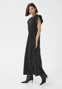 Črna maksi obleka z nabranimi rokavi in elegantno silhueto. Model jo kombinira z črnimi čevlji na peto v obliki konice za sodoben videz.