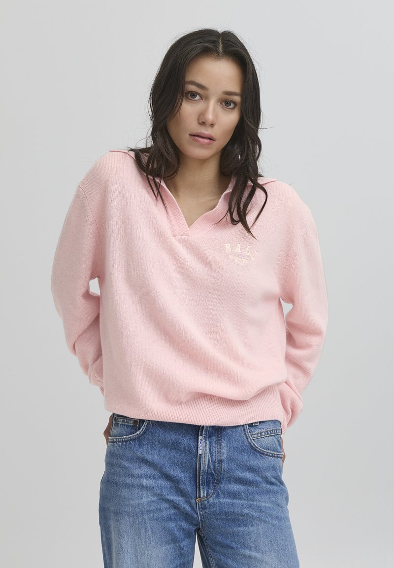 Ružičasti pletivi džemper s V-izrezom, s diskretnim logotipom i rebrastim manžetama, u kombinaciji s plavim trapericama. Jednostavan i moderan stil.