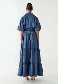 Modrá dlouhá maxi šaty s vrstveným střihem, nabíranými rukávy a uvázaným pasem. Vyrobeny z měkké látky, s volným střihem a volánkovým lemem.