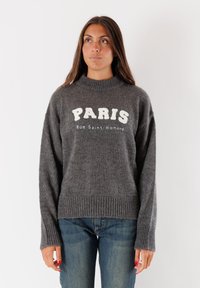Donna con lunghi capelli castani che indossa un maglione grigio lavorato a maglia con la scritta "PARIS Rue Saint-Honoré", abbinato a jeans blu, in piedi su uno sfondo bianco.