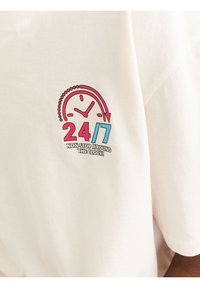 Lys beige bomulds T-shirt med et farverigt graphic: et ur med røde og blå accenter, tekst "24/7 NON STOP RUNDT OM URET!"