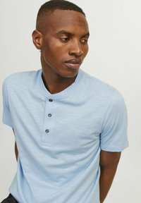 Polo de manga corta azul claro con cuello redondo, que presenta tres botones grises y un acabado sutilmente texturizado.