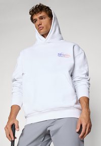 White hoodie in cotton blend, featuring a front kangaroo pocket and colorful "Students Social Studies" graphic on the chest.