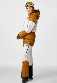 Ski suit with a brown and white color scheme, featuring zippers, pockets, and a hood. Made from textured, waterproof fabric.