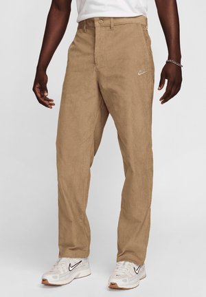 Mann trägt beige Nike-Cordhosen und weiße Nike-Sneaker, steht mit entspannten Armen an den Seiten vor einem hellgrauen Hintergrund.