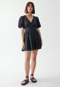 Schwarzes kurzes Kleid mit Puffärmeln, tiefem V-Ausschnitt, taillierter Passform und gerafftem Rock. Model trägt schwarze Riemchensandalen.