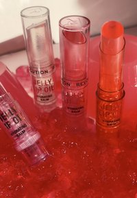 Fire cylinders med leppebalsam i klart og rosa, merket "Jelly Lip Oil" og "Hydrating Balm," plassert på en livlig rød gel bakgrunn.