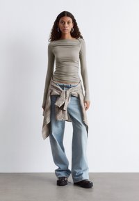 Langärmliges, olivgrünes, figurbetontes Oberteil mit seitlichem Raffung, kombiniert mit lockeren, hellblauen Jeans und einem gebundenen Karohemd in Beige- und Rosatönen. Schwarze Plateauschuhe.