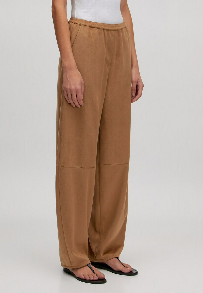 Beige wijde broek gemaakt van zacht materiaal met een elastische tailleband, voorzien van naaddetails en een relaxte pasvorm, gecombineerd met platte sandalen.