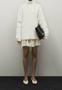 Pull en maille blanc surdimensionné avec col haut, associé à une jupe courte crème. Un sac à main noir et des ballerines blanches pointues complètent la tenue.