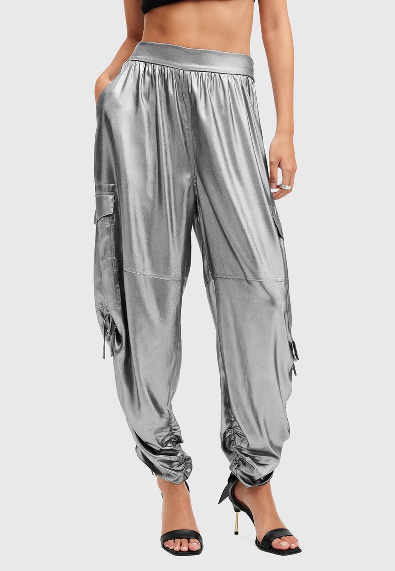 Femme portant un pantalon cargo argenté brillant avec des poignets élastiques et des poches, associé à des sandales à talons hauts noires et un top court noir.