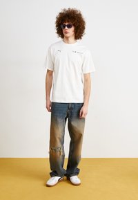 Camiseta blanca de algodón con logo, vaqueros de mezclilla azul desgastados, degradado de claro a oscuro, y zapatillas blancas con suelas marrones.