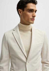 Blazer blanc à rayures avec revers crantés, fermeture à un bouton et poche poitrine, superposé à un pull à col roulé beige.