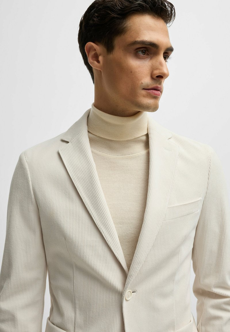 Witte geribbelde blazer met notch-lapels, enkele knoopsluiting en een borstzak, gelaagd over een beige coltrui.