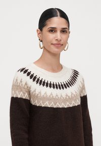 Kvinne med strammet mørkt hår som har store gulldobbelte øreringer og en brun, beige og hvit mønstret ullgenser mot en ensfarget bakgrunn.