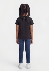 Παιδί με κοτσίδες, φορώντας μαύρο κοντομάνικο μπλουζάκι, μπλε τζιν και άσπρα αθλητικά παπούτσια, στέκεται με την πλάτη προς τα εμπρός σε απλό φόντο.