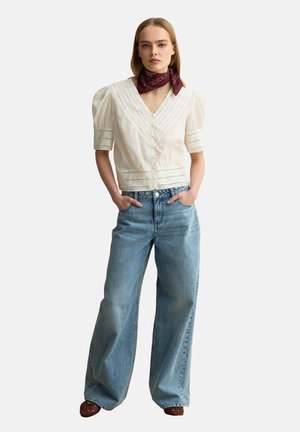 Mujer con blusa blanca con encaje, vaqueros azul claro y holgados, zapatos marrones y un pañuelo burdeos con estampado en el cuello, de pie con las manos en los bolsillos.