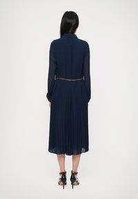 Γυναίκα φορώντας έν�α ναυτικό μπλε πλισέ μίντι φόρεμα με μακριά διάφανα μανίκια, μια χρυσή ζώνη αλυσίδα και μαύρα ψηλοτάκουνα πέδιλα με λουράκι στον αστράγαλο, σε θέα από πίσω.