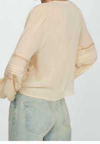 Blusa beige con maniche ampie, dettagli plissettati e accentuazioni in pizzo, indossata su jeans chiari. Tessuto morbido e leggero.