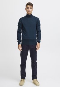 Jūrine turtlenecko tipo megztinis su tekstūriniu raštu, derinamas su tamsiomis kelnėmis ir smėlio spalvos batais. Modelis stovi prieš paprastą baltą foną.