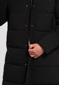 Abrigo acolchado negro con cierre frontal, botones a presión y grandes secciones acolchadas. Cuenta con un cuello alto y bolsillos laterales con cremallera.