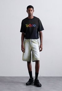 Chemise noire à manches courtes avec le texte "Techno" multicolore, associée à un short gris à motifs. Des chaussettes noires et des baskets complètent la tenue.