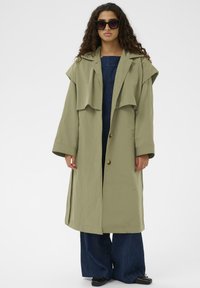 Olivnozelen plašč dolgega kroja z spuščenimi rameni, zapiranjem na gumbe in širokimi rokavi. V kombinaciji s temnimi jeansi širokega kroja.