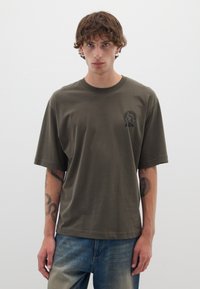 Camiseta de algodón verde oliva de manga corta, con cuello redondo y un pequeño logotipo gráfico negro en el lado izquierdo del pecho.