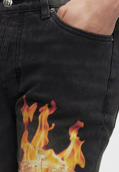 Χέρι στην τσέπη μαύρου τζιν με πορτοκαλί και κίτρινο σχέδιο φλόγας κοντά στον μηρό σε λευκό φόντο.
