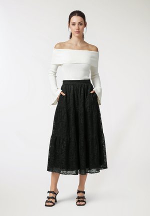 Donna in piedi, indossa un top bianco a coste con scollo a barchetta, una gonna midi nera in pizzo con tasche e sandali neri con tacco e fiocchi.