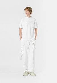 Camiseta blanca de gran tamaño con un logo texturizado, combinada con pantalones jogger blancos que cuentan con bolsillos laterales y detalles de costura en contraste. Calzado en tono pálido.