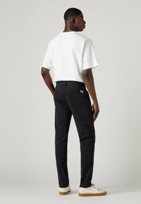 Black pants with a straight-leg design, paired with a white t-shirt. Textured fabric, two back pockets, and subtle branding detail visible.