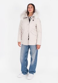 Parka de color beige claro con capucha de piel sintética, presenta botones a presión, dos bolsillos frontales y un corte relajado. Combinada con jeans azules y zapatillas blancas.