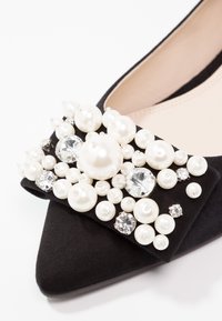 Chaussure plate noire à bout pointu avec un nœud décoratif orné de perles blanches de différentes tailles et de strass transparents.