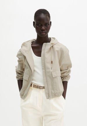 Femme à la peau foncée portant une veste beige à capuche et fermeture éclair sur un débardeur blanc avec un pantalon taille haute crème et une ceinture beige.