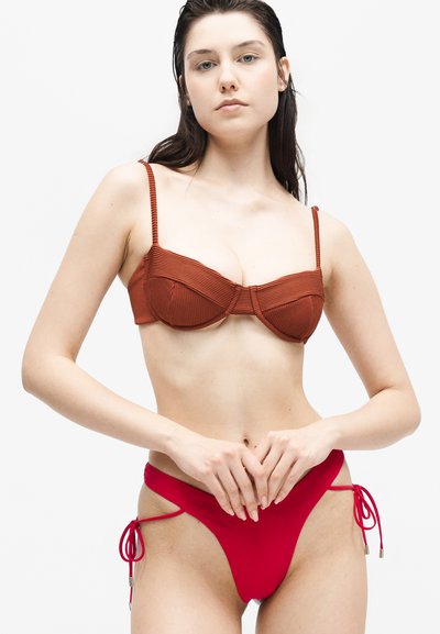 Νέα γυναίκα με μακριά σκούρα μαλλιά που φοράει ένα σκουροκόκκινο ριμπ κορμάκι μπικίνι και κόκκινα κάτω μέρη μπικίνι με κορδόνια στα πλάγια, στέκεται μπροστά σε ένα λευκό φόντο.