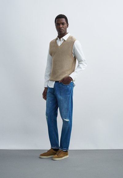 Νέος άνδρας στέκεται με το ένα χέρι στην τσέπη, φοράει μπεζ πλεκτό γιλέκο πάνω από λευκό πουκάμισο, μπλε ripped τζιν και καφέ αθλητικά παπούτσια σε γκρι πάτωμα.