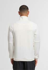 Valge tõmblukuga sviiter kõrge kaelusega, millel on tekstureeritud kangas ja reljeefne brändilogo selja peal. Pikkade varrukatega, millel on mansetid.