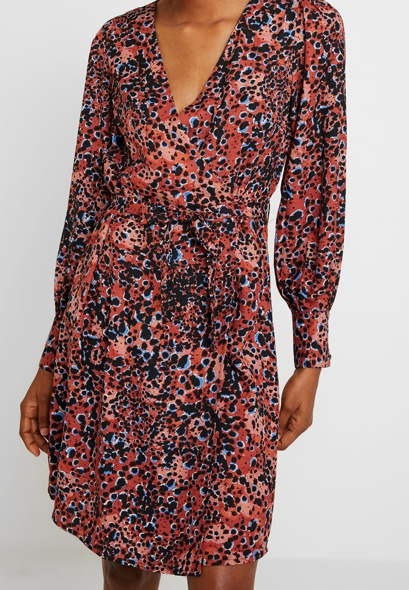 Femme portant une robe portefeuille à manches longues avec une ceinture à la taille, présentant un motif abstrait à taches rouges, noires et bleues.