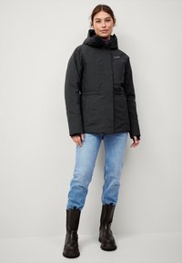 Zwarte geïsoleerde jas met een capuchon, verticale naaddetails, zijzakken, gedragen over een paarse top, gecombineerd met lichtblauwe jeans en zwarte laarzen.