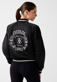 Giacca bomber nera con logo ricamato bianco e motivo floreale sulla schiena, polsini a coste e design sportivo con vestibilità comoda.