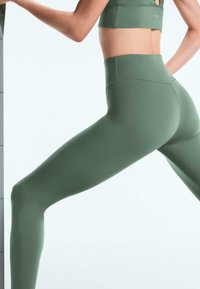 Oseba v zelenih visokih pajkicah in ujemajočem športnem nedrčku razteza eno nogo ob steni ali površini.