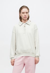 Pulover gri deschis cu guler și țesătură texturată. Prezintă o deschidere cu nasturi și manșete striate, asortat cu pantaloni de trening roz.