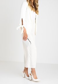 Blazer blanco con mangas atadas, combinado con pantalones de pierna ancha a rayas claras. Tacones cerrados blancos y un clutch plateado completan el atuendo.