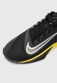 Chaussure de sport noire avec une tige en tissu texturé, logo swoosh blanc, accents jaunes sur la semelle et une semelle en caoutchouc à motif.
