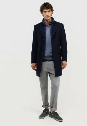 Hombre con abrigo azul marino, suéter azul, camisa burdeos, pantalones gris claro remangados en los puños y zapatos casuales grises, de pie frente a un fondo liso.